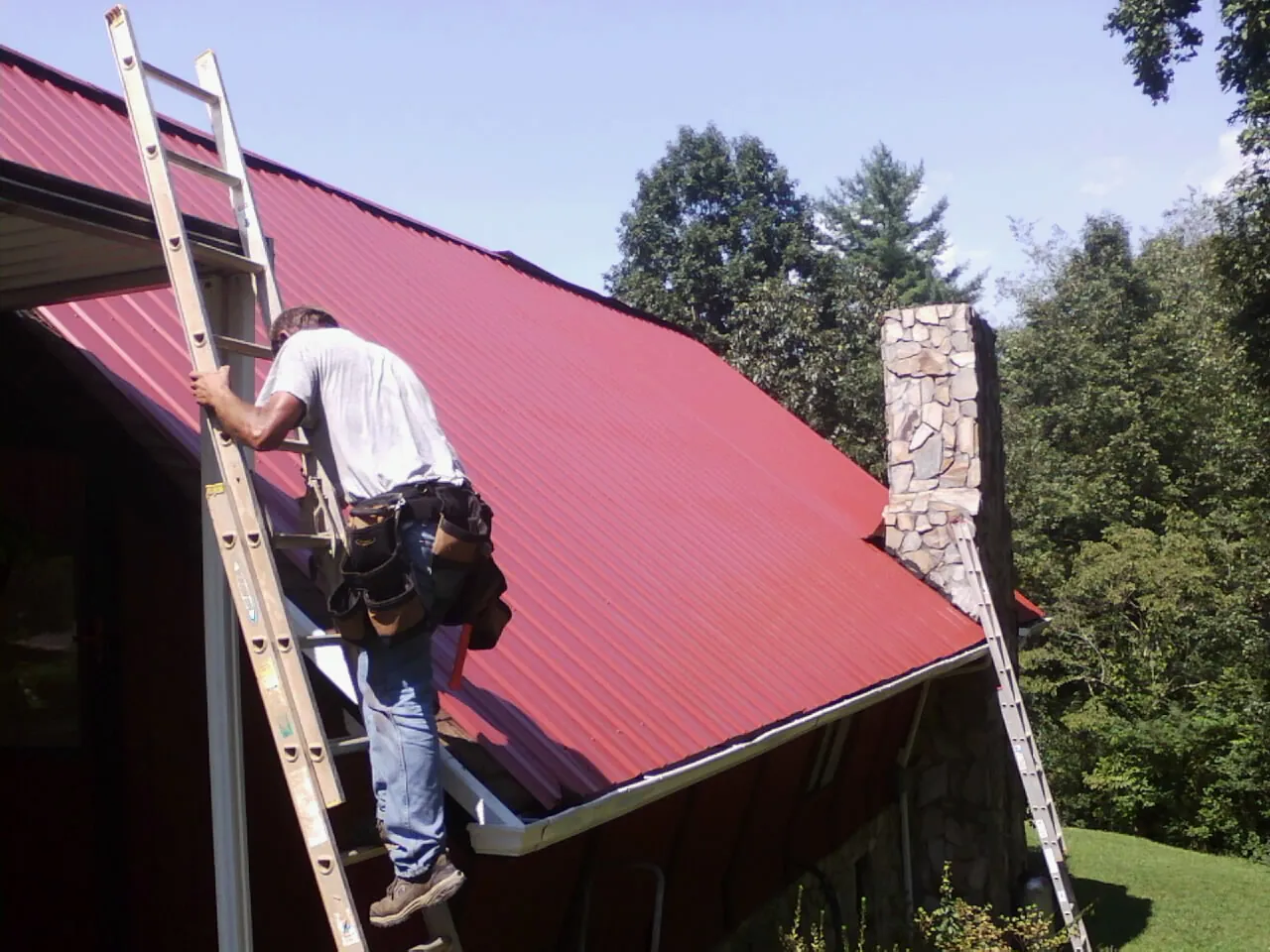 Roofer on a ladder beside a red metal roof and stone chimney — Soffit & Fascia Repair by Roofing Brothers in Locke